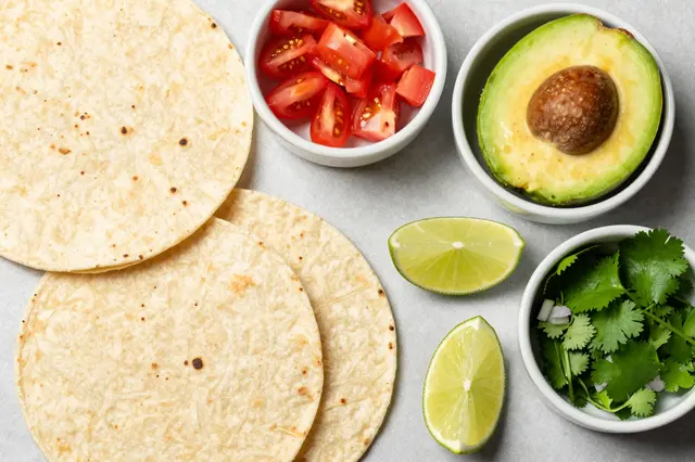 Top-down flat lay of corn tortillas and fresh taco toppings arranged in small bowls on a light gray countertop