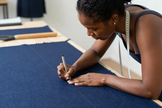 A female tailor with a measuring tape around her neck marks lines on navy wool fabric at a cutting table.