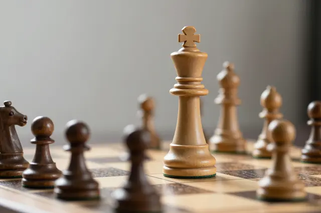 Focused light wooden chess king stands tall surrounded by darker opposing pieces on a chess board with blurred neutral