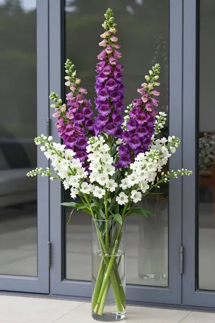 A tall glass vase of purple foxgloves and white blooms sits beside gray framed glass garden doors.