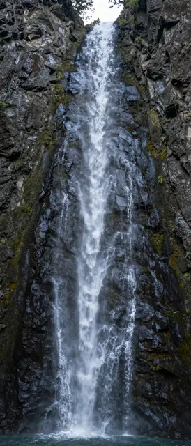 A slender cascading waterfall flows down a steep dark rocky gorge into a small pool below