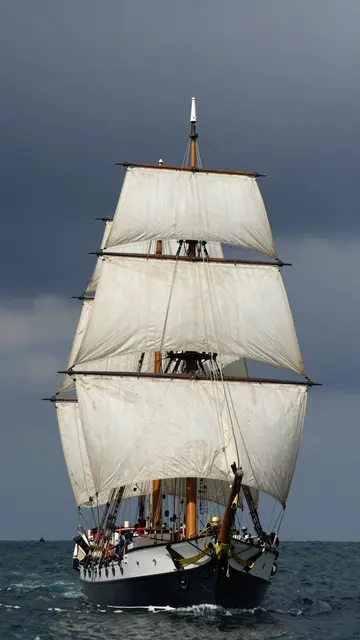 A tall sailing ship with full billowed sails moves across dark choppy sea under a heavy stormy overcast sky.