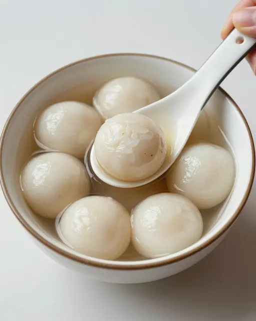 A bowl of glutinous rice balls in light syrup, with a porcelain spoon held by a partial hand lifting one tangyuan.