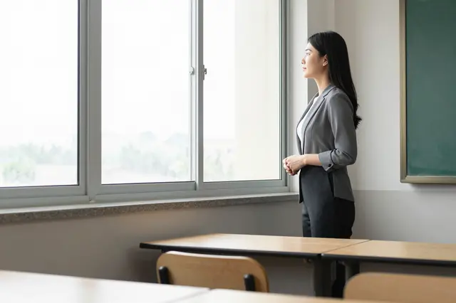 A side profile view of a female teacher pausing while looking out a window in an empty classroom