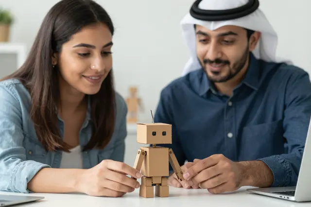 Two smiling colleagues examine a small cardboard robot prototype on an office table