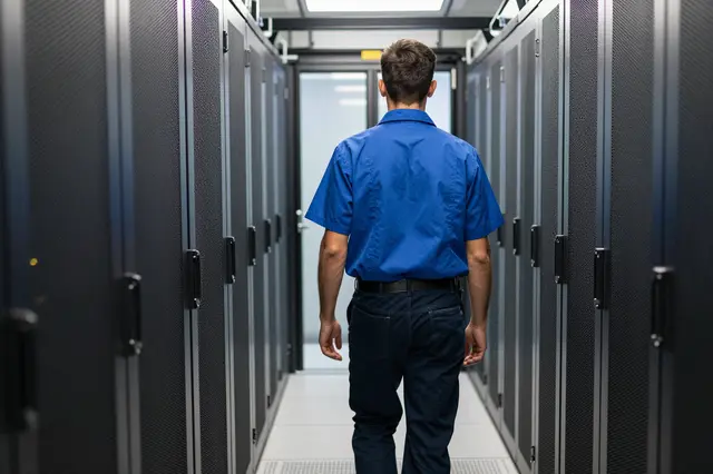 Viewed from behind, a technician in a blue shirt walks down a narrow data center server aisle.