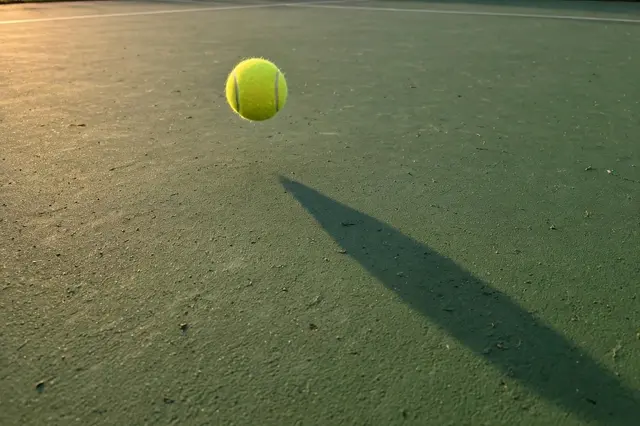 A yellow tennis ball hovers mid-air above a green tennis court, casting a long evening shadow.