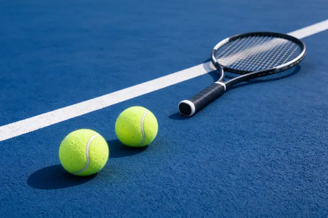 Two yellow tennis balls and a tennis racket rest on a marked blue hard tennis court in bright sunlight.