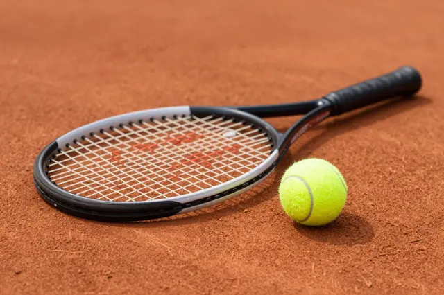 A tennis racket and a bright yellow tennis ball rest on a textured reddish clay tennis court.