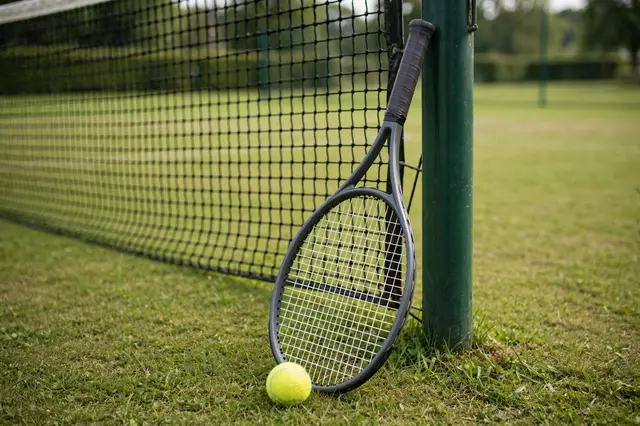 A tennis racket leans against a net post with a yellow tennis ball at its base on a grass tennis court.