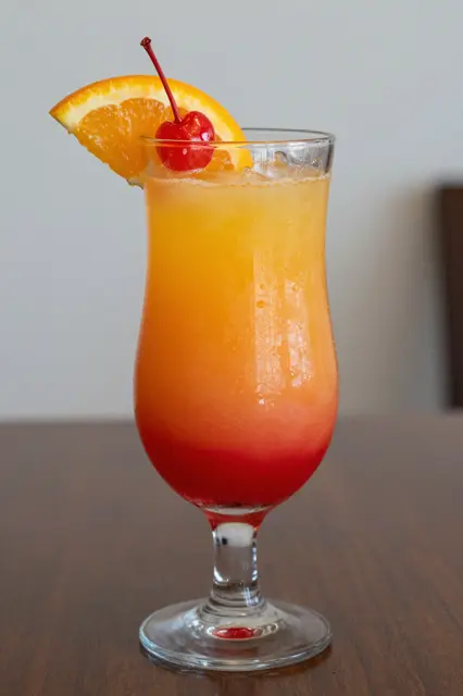 A tall stemmed glass holding a gradient tequila sunrise garnished with an orange slice and maraschino cherry.