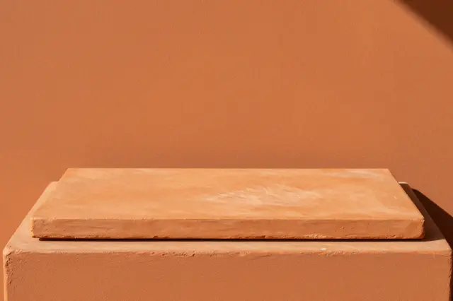 Terracotta display plinth sits against a burnt orange wall in warm side sunlight with copy space
