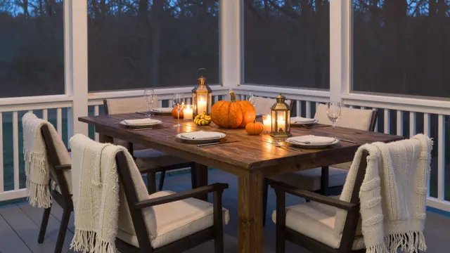 Set wooden Thanksgiving dining table on a screened porch at dusk with lanterns, pumpkins and knit blankets draped over chairs