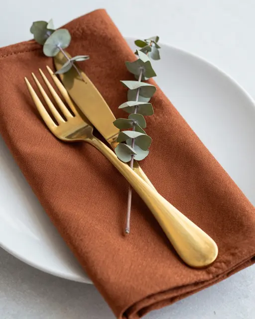 A styled Thanksgiving place setting with gold cutlery, rust napkin, eucalyptus sprig on a white plate