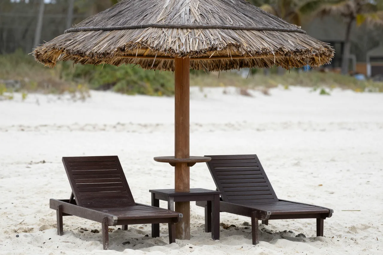 Two dark wooden sun loungers sit under a straw thatched umbrella on a quiet white sand beach