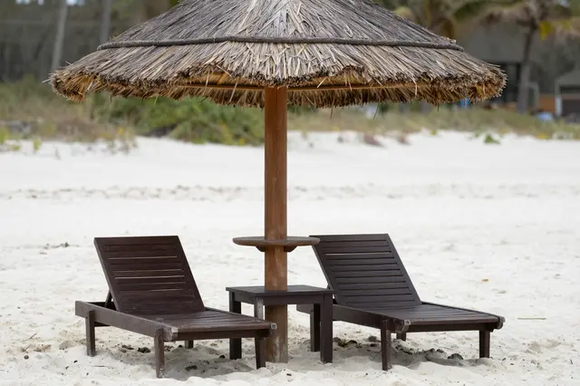 Two dark wooden sun loungers sit under a straw thatched umbrella on a quiet white sand beach
