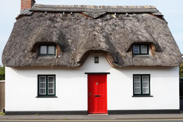 A white traditional thatched cottage with a bright red front door set beside a quiet street