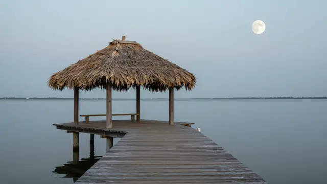 Wooden jetty leading to a thatched shelter over calm lagoon beneath a bright full moon at twilight
