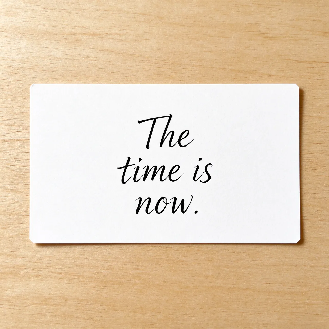 A white index card on a light wooden desk displays the text "The time is now." in black ink