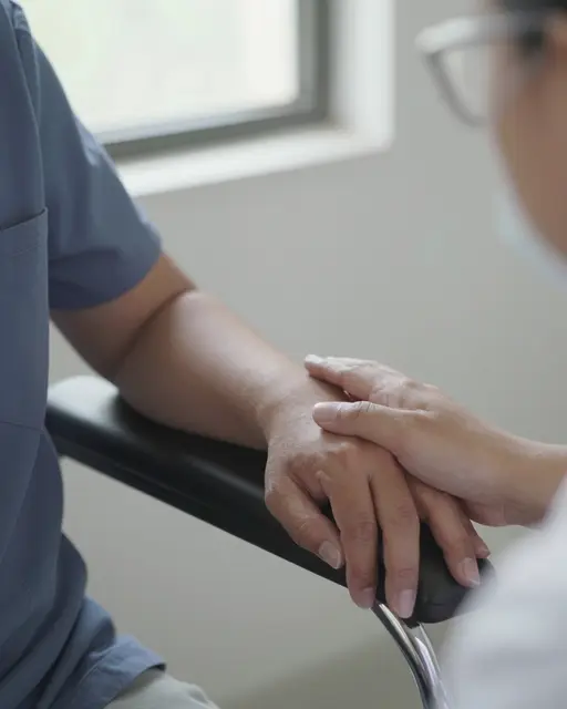 A healthcare provider rests a gentle hand over a patient's hands on a chair arm in soft window light