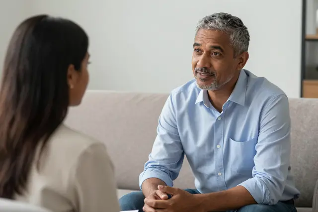 A middle aged male counselor listens attentively to a South Asian woman during an office therapy session