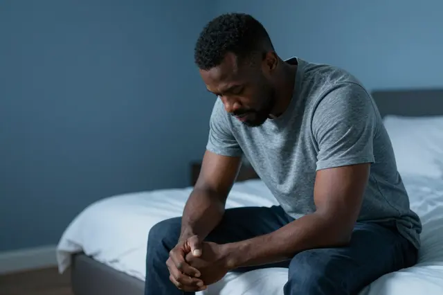 A Black man in his forties sits bowed with clasped hands on the edge of a bed in dim blue early morning light