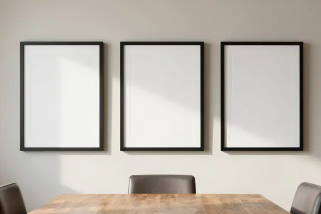 Three empty black picture frames hang above a wooden dining table in soft daylight.