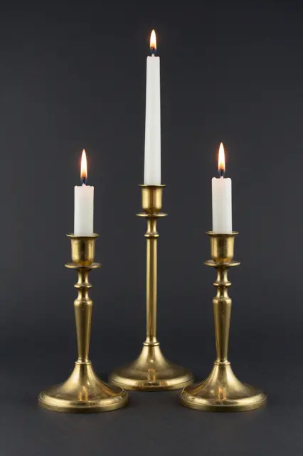 Three varying height brass candlesticks with lit white tapers against a dark background