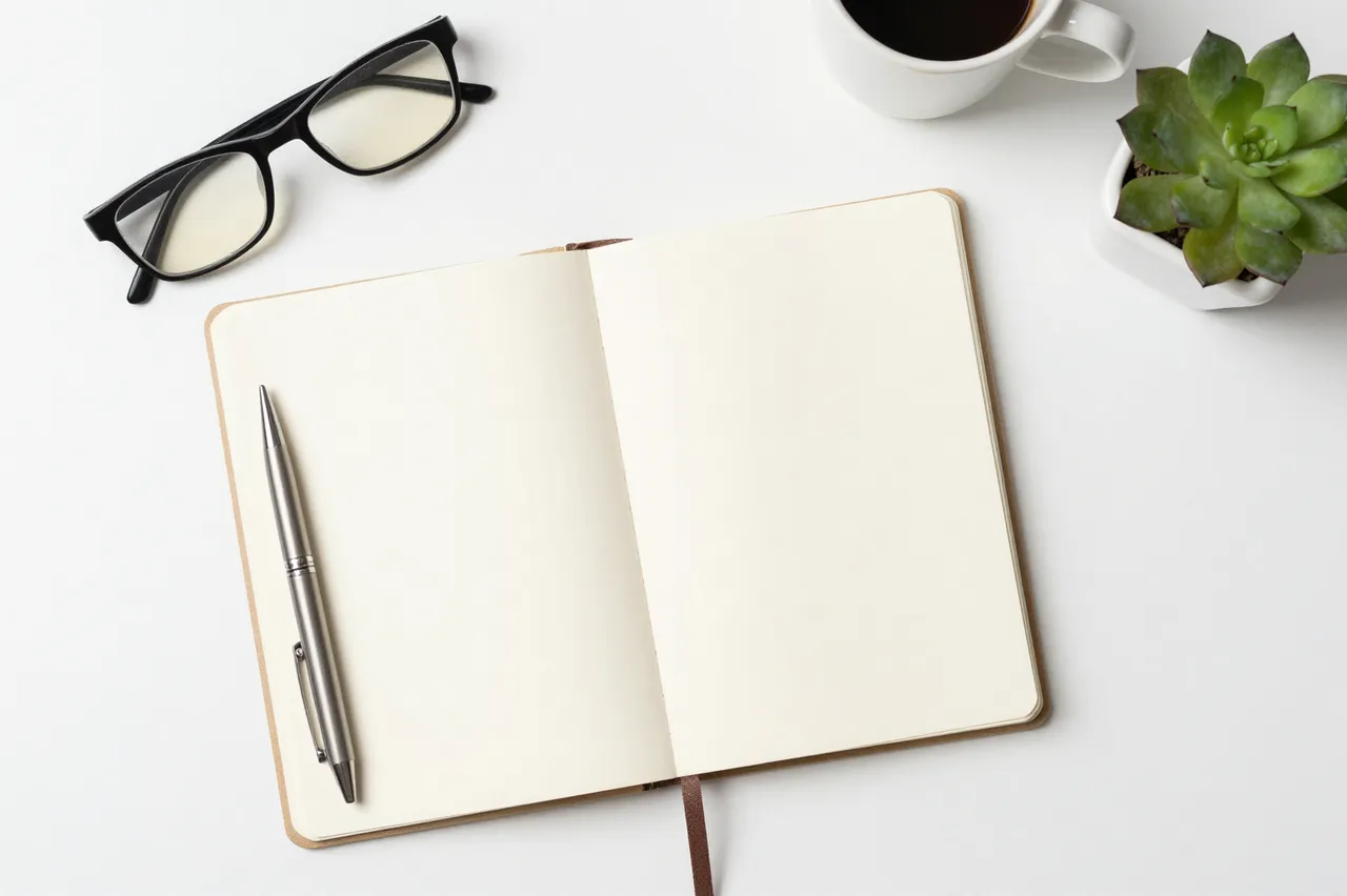 Top-down view of an open blank notebook, pen, eyeglasses, coffee cup and succulent on a white desk surface