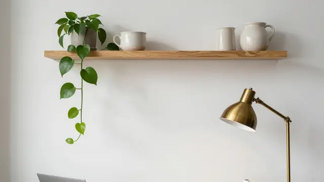 A wall-mounted oak shelf holds plain ceramics and a pothos next to a brass desk lamp on a neutral home office wall.