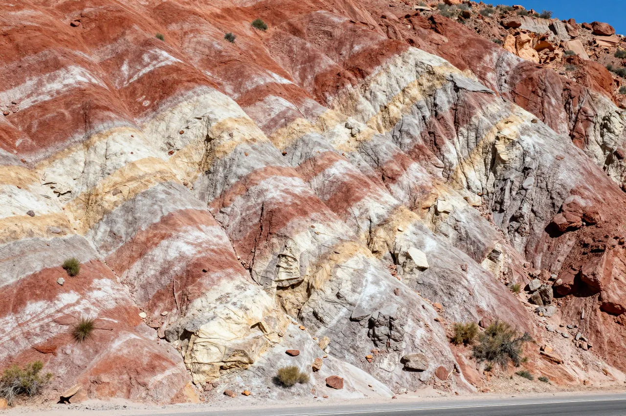 Exposed tilted layers of red, cream and gray sedimentary rock line a roadside under bright midday sun