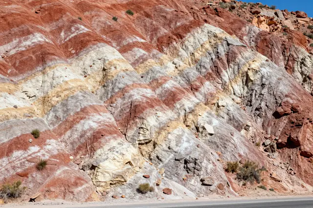Exposed tilted layers of red, cream and gray sedimentary rock line a roadside under bright midday sun