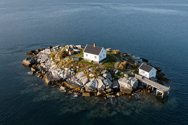 Aerial view of a small rocky ocean island with white buildings, a wooden jetty, and surrounding deep blue water.