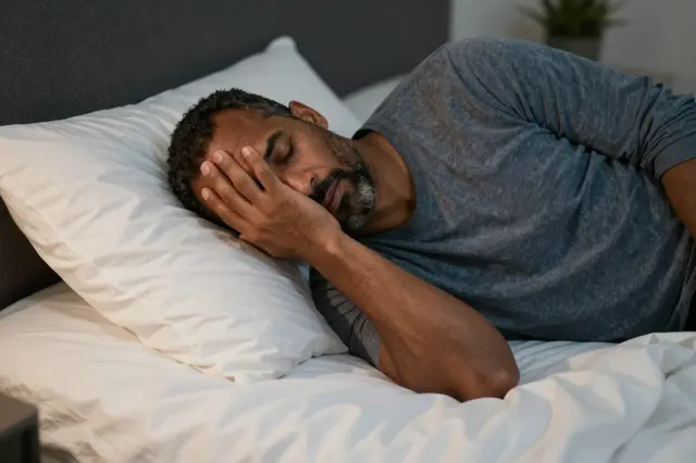 A tired mixed-race man in his forties rests on a bed with his hand over his eyes in dim evening light.