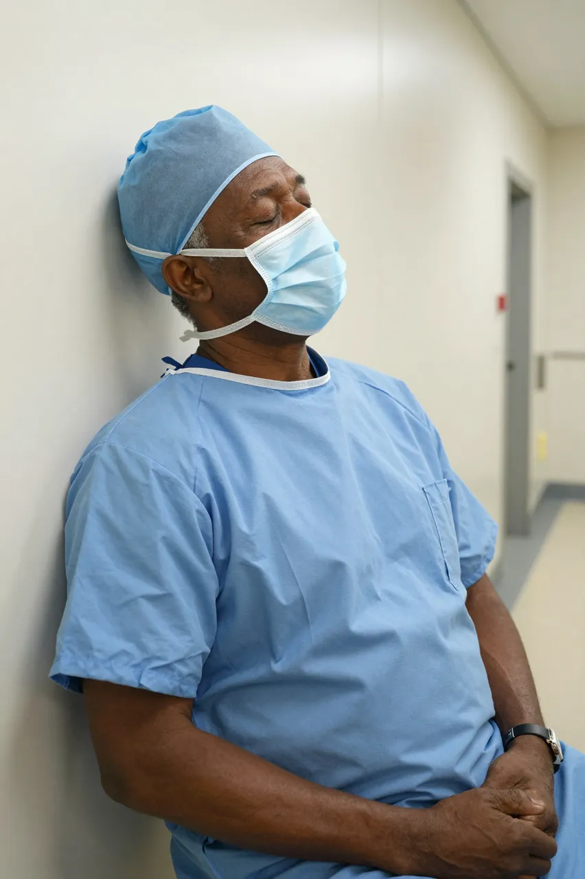 Older Black surgeon in blue scrubs leans against a hospital corridor wall with eyes closed, surgical mask in place over his