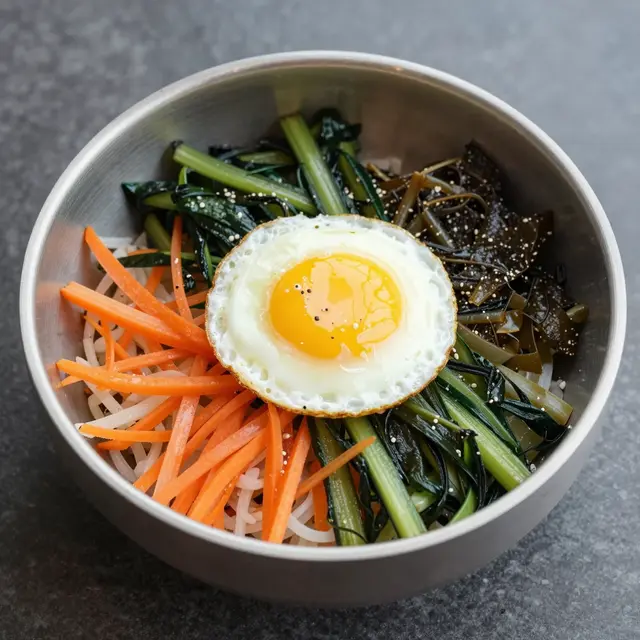 A top-down view of a metal bibimbap bowl with assorted vegetables and a centered fried egg on a gray countertop.