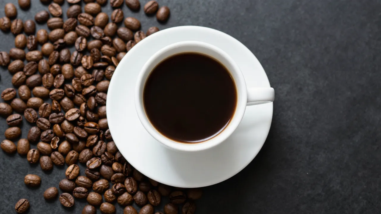 A top-down view of a white cup of black coffee resting on a saucer next to scattered roasted coffee beans on a dark stone