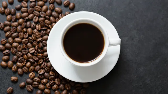 A top-down view of a white cup of black coffee resting on a saucer next to scattered roasted coffee beans on a dark stone
