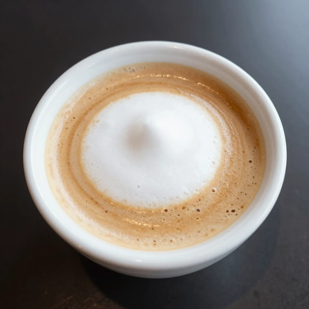 Overhead close-up of an espresso macchiato with a foam dollop in a small white cup on a dark table.