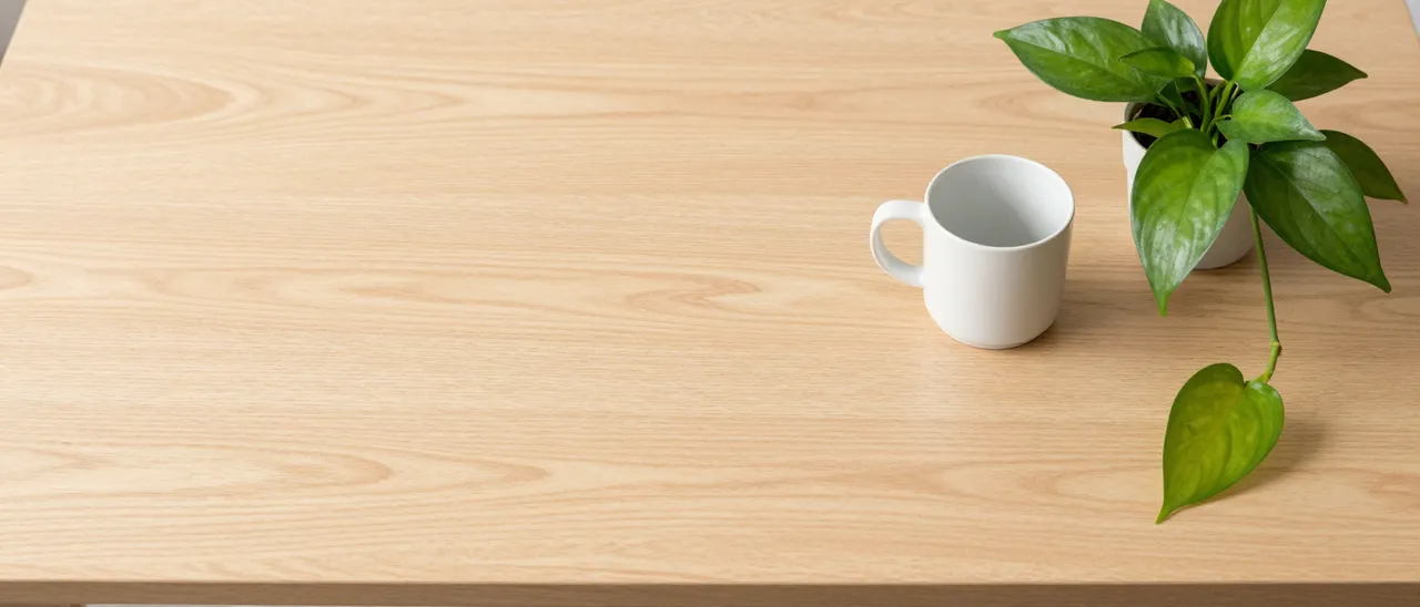 Overhead view of a pale oak desk with empty left space, a white ceramic mug, and a trailing pothos plant on the right.