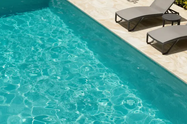 A high angle view of a turquoise swimming pool with two grey lounge chairs on a beige tiled patio