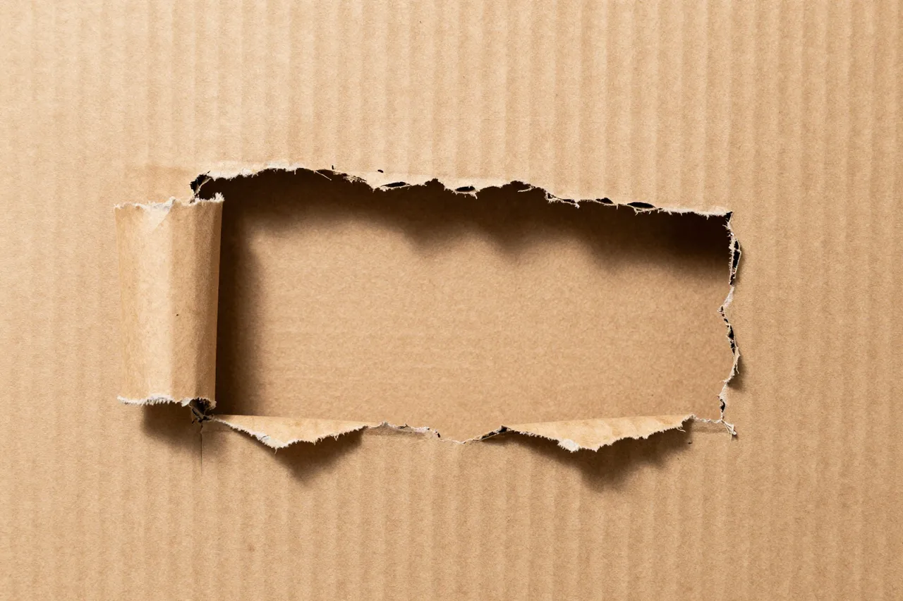 Close-up of torn brown corrugated cardboard with a ragged rectangular opening and curled left edge