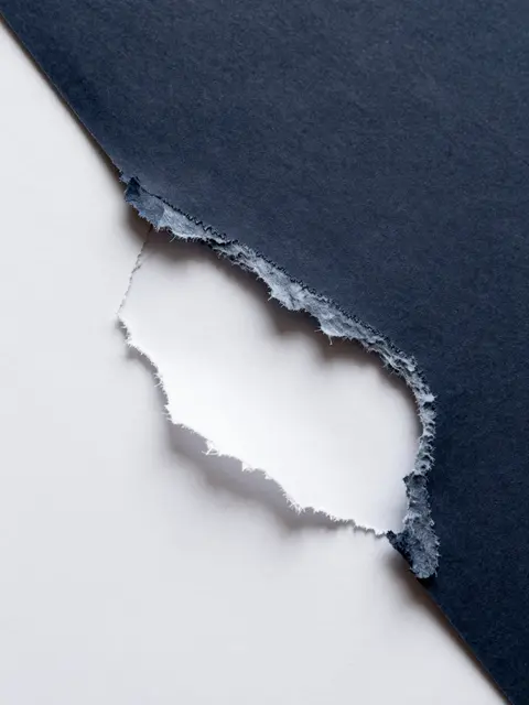Close-up of frayed torn dark navy paper resting atop a plain white sheet under flat even lighting