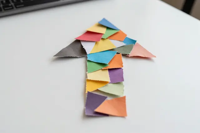An upward arrow assembled from torn multicolored paper rests on a white office desk