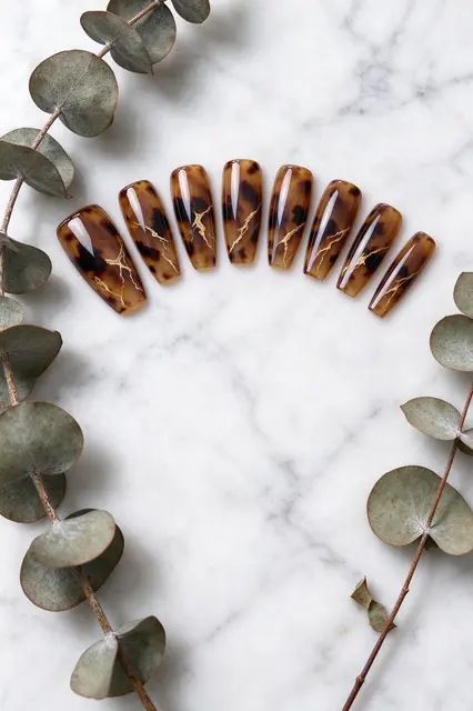 Glossy tortoiseshell press-on nails arranged in an arc with dried eucalyptus on white marble