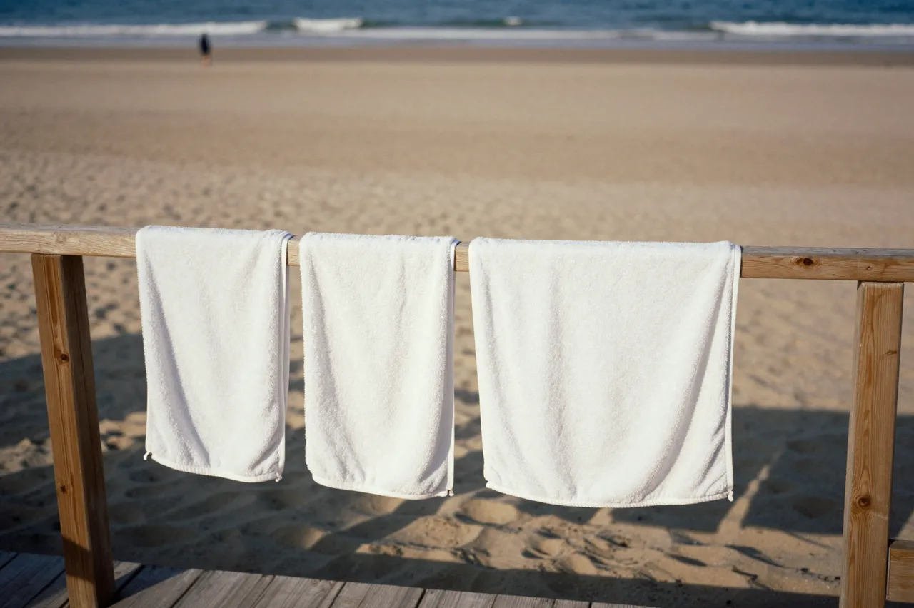 Three white terry towels hang on a wooden railing overlooking a wide sandy ocean beach in warm late afternoon light.