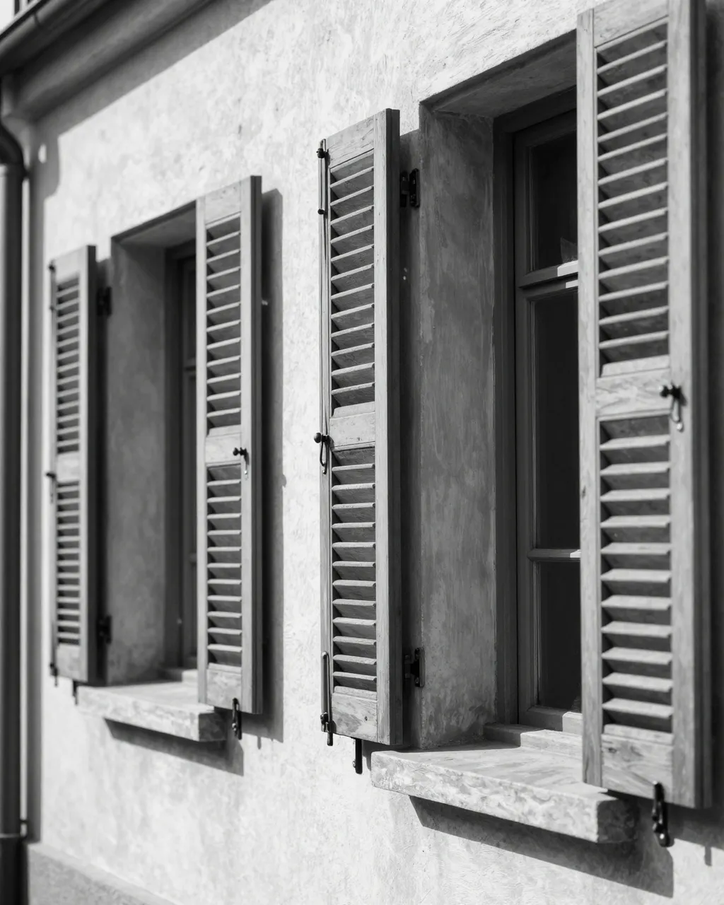 Black and white view of half-open wooden louvered shutters on an old townhouse wall with slanted sunlight