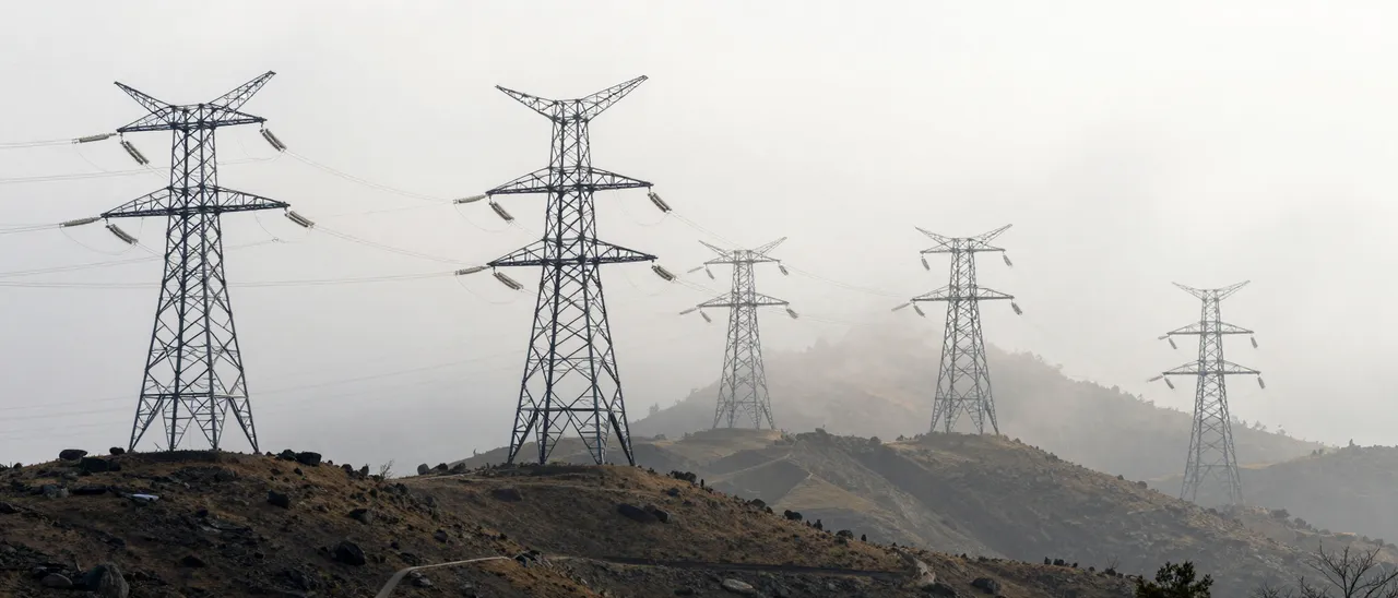 A line of electricity transmission towers stands in a receding row across rocky fog-shrouded hills