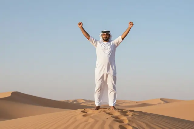 Middle Eastern man in traditional clothing stands on a sand dune crest with raised fists at sunrise