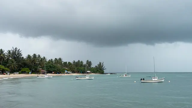 Small anchored boats rest on calm turquoise bay water under a heavy gray storm sky beside a palm-lined beach